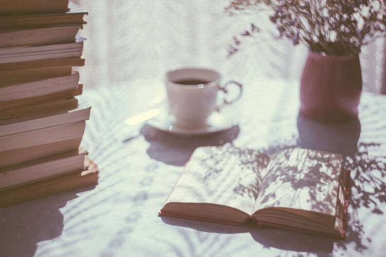 A cozy scene featuring a stack of books, an open book, a cup of coffee on a saucer, and a vase with flowers on a sunlit table.