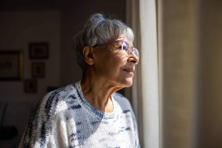 A senior woman, partly in shadow, gazes through a window to the outside. (iStock/adamkaz)