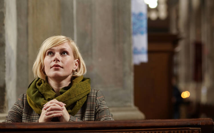 A young woman kneels and prays at a pew, looking toward the altar of a Catholic church. (iStock/roman_sh)