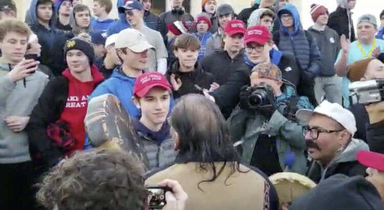 On Jan. 18, a teenager wearing a "Make America Great Again" hat, center left, stands in front of an elderly Native American singing and playing a drum in Washington. (Survival Media Agency via AP)