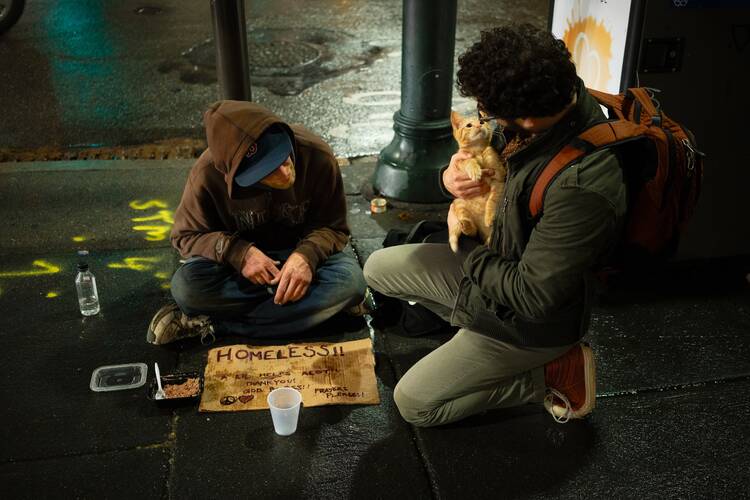 a man is seated on the ground with a cardboard sign that reads "homeless please help" and a man crouches holding a kitten next to him