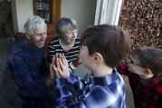Two grandparents, an older white woman and white man with grey hair, sit behind a glass or plastic screen, touching hands with two of their grandchildren, a boy and a girl, who from their profile are also light-skinned.
