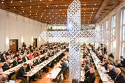 A metallic, contemporary-styled cross in the foreground while German bishops sit at long tables in the background.
