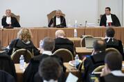 The judges of the Vatican City State criminal court—Venerando Marano, Giuseppe Pignatone and Carlo Bonzano—sit at a long table with a brown tablecloth in a makeshift court room.