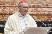 A man in white robes wearing the red cap of a cardinal speaks into a microphone