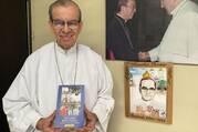 cardinal gregorio rosa chavez holds a copy of a book with a photo of oscar romero and a photo of oscar romero with the pope behind him