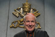 cardinal christoph schönborn sits wearing his black clothes and red cap with the papal keys seal behind him