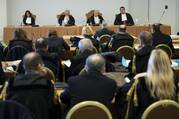 four men sit at a table in front of a large crowd in a makeshift courtroom in the vatican museums