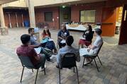 seven young people sit around in chairs talking to each other in a courtyard area inside a building