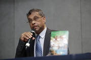 aloysius john wears a suit in front of a dark wall and speaks into a microphone. he is a french man of indian descent