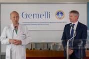 dr sergio alfieri wearing a white labcoat speaks next to matteo bruni who wears a navy suit. they stand in front of a sign that reads "gemelli hospital"