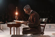 The actor Jeremy Strong sitting at a desk reading a book by candlelight in a theatrical production of the play Enemy of the People