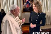 This image shows Pope Francis conversing with a woman in an elegantly decorated room. Pope Francis, dressed in his traditional white papal attire, appears to be speaking while gesturing with his hand. The woman, wearing a black dress adorned with golden buttons and puff shoulders, is smiling warmly as she listens. Behind them, a large painting featuring a religious scene enhances the formal setting. The image has a watermark of “CBS News” in the bottom right corner.