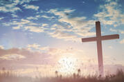 A cross in silhouette against the sunset in a field with birds flying in the distance.