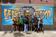 seven college students in regular clothes stand in front of a graffiti mural of george floyd with his face and name, in minneapolis