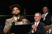 A man in a tan coat and black tie holds a trophy