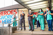 health care workers clapping outside a hospital wearing masks during covid, there is a sign behind them denoting them as heroes