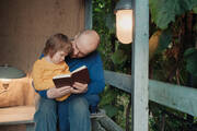 A man reads to a toddler sitting on his lap on a porch