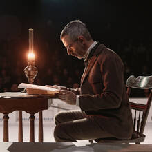 The actor Jeremy Strong sitting at a desk reading a book by candlelight in a theatrical production of the play Enemy of the People