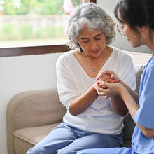 A young female doctor in blue scrubs holds hands with an older female patient, both sitting on a couch. (iStock/BongkarnThanyakij)
