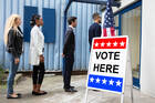Young men and women stand in line outside a polling place with a "Vote Here" sign.